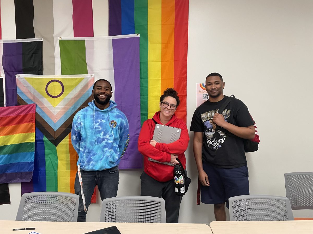 (From left to right) Jordain Johnson, Esmeralda Darosa and Tyler Watson smiling, standing in front of pride flags in the Multicultural Center.